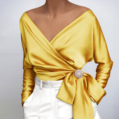 Blouse en satin jaune à manches longues, col en V, nouée à la taille avec un nœud élégant, portée avec un pantalon blanc. Mode féminine chic.