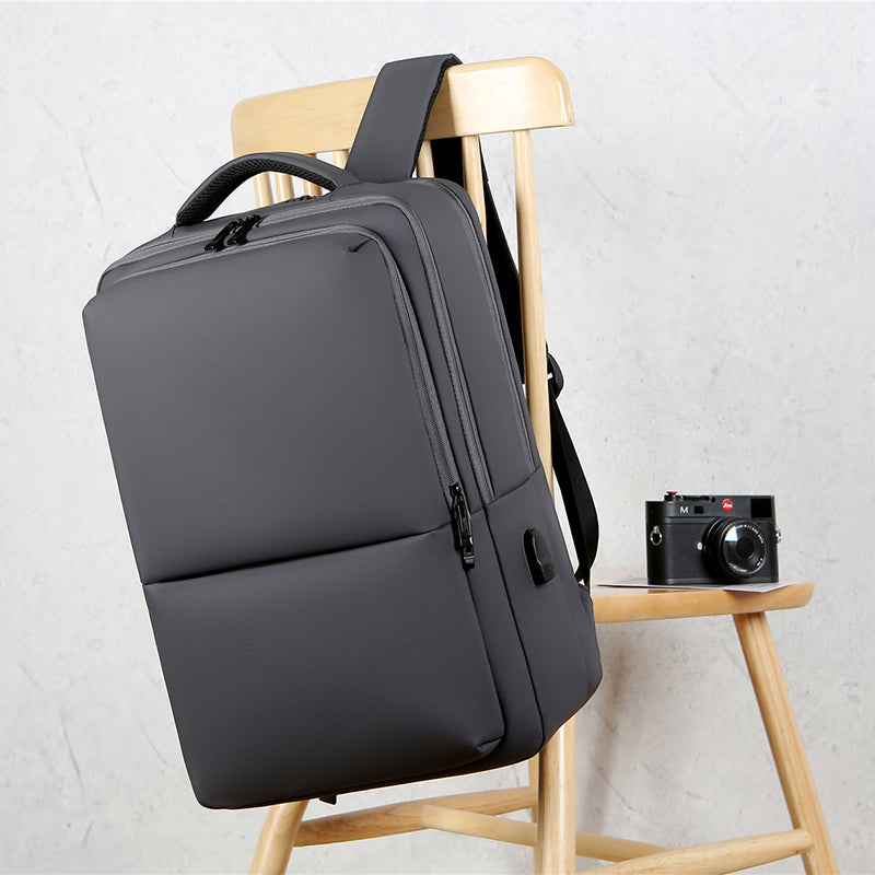 Sac à dos gris moderne sur chaise en bois, à côté d'un appareil photo vintage. Design minimaliste, idéal pour le voyage et le quotidien.