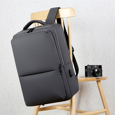 Sac à dos gris moderne sur chaise en bois, à côté d'un appareil photo vintage. Design minimaliste, idéal pour le voyage et le quotidien.