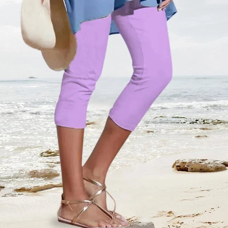 Femme en pantacourt violet sur plage, portant sandales dorées et chapeau, ciel bleu en arrière-plan. Mode estivale, tenue décontractée bord de mer.