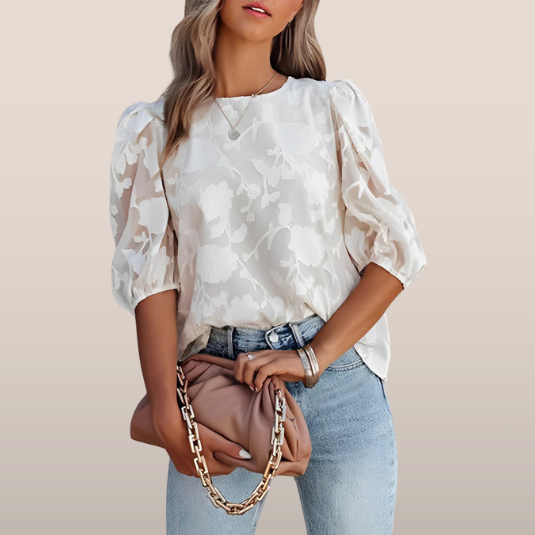 Femme portant un chemisier blanc à motifs floraux, manches bouffantes, avec un sac à main beige à chaîne dorée, posant en jean bleu clair.
