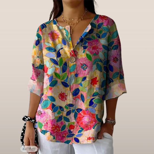 Blouse femme à manches trois-quarts, motif floral coloré, col en V, portée avec un collier doré et un pantalon blanc. Mode printemps-été tendance.