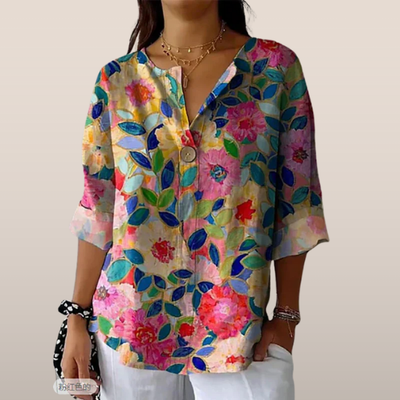 Blouse femme à manches trois-quarts, motif floral coloré, col en V, portée avec un collier doré et un pantalon blanc. Mode printemps-été tendance.