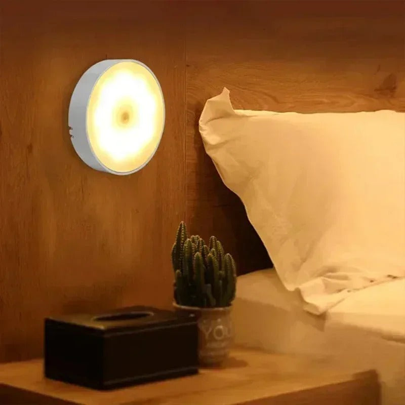 Lampe murale LED ronde émettant une lumière chaude, installée dans une chambre moderne avec lit et table de chevet en bois, ambiance cosy.