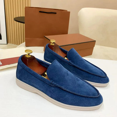 Mocassins en daim bleu pour homme avec embauchoirs dorés, posés sur une table blanche, sac et boîte en arrière-plan. Chaussures élégantes et modernes.