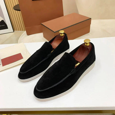Paire de mocassins noirs en daim pour homme sur une table blanche, semelle blanche, embauchoirs dorés, boîte à chaussures marron en arrière-plan.