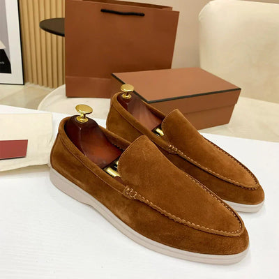 Mocassins en daim marron pour homme avec embauchoirs dorés, posés sur une table blanche, boîte et sac en arrière-plan. Chaussures élégantes et modernes.
