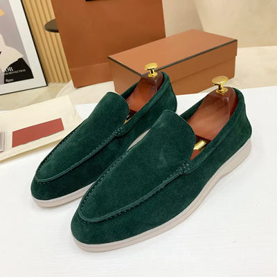 Mocassins en daim vert foncé élégants avec embauchoirs dorés, posés sur une surface beige, boîte marron en arrière-plan. Chaussures homme tendance.