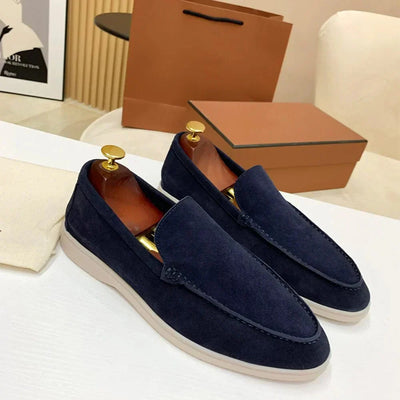 Mocassins en daim bleu marine pour homme sur une table blanche, avec embauchoirs dorés, boîte à chaussures marron en arrière-plan. Chaussures élégantes.