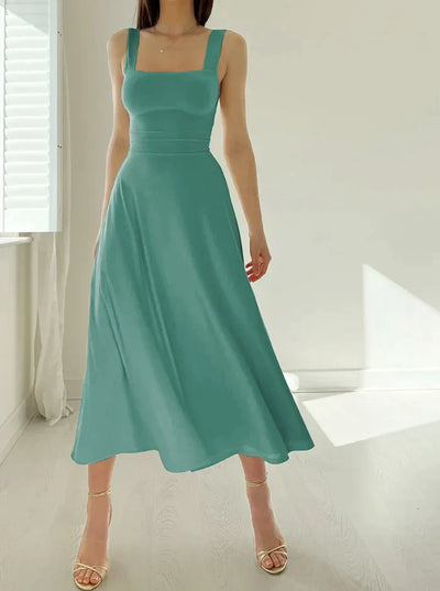 Robe midi verte élégante sans manches, portée par une femme, dans une pièce lumineuse avec parquet clair et fenêtre à persiennes blanches.