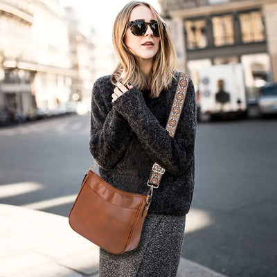 Femme portant un sac à bandoulière en cuir marron, pull gris, jupe, lunettes de soleil, rue urbaine. Mode automne, accessoires tendance.