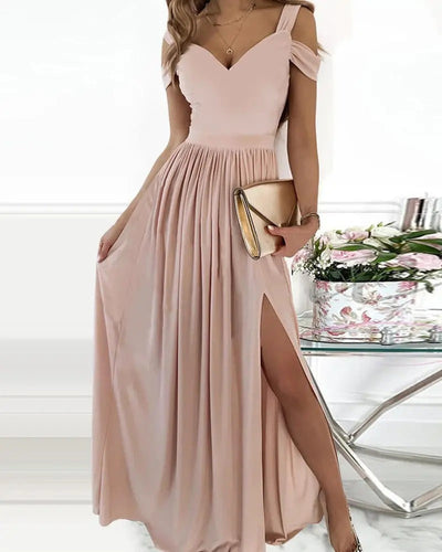 Robe longue rose pâle élégante avec fente, tenue de soirée chic, femme tenant une pochette dorée, décor floral en arrière-plan.