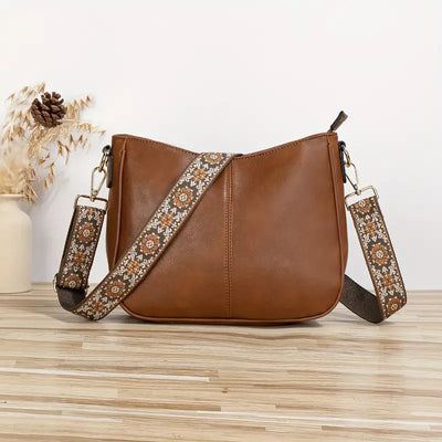 Sac à main en cuir marron avec bandoulière à motifs floraux, posé sur une table en bois clair, décoré de fleurs séchées en arrière-plan.