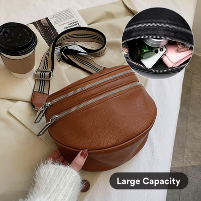 Sac banane en cuir marron avec fermetures éclair argentées, grande capacité. Accessoires visibles: téléphone, écouteurs. Tasse de café en arrière-plan.