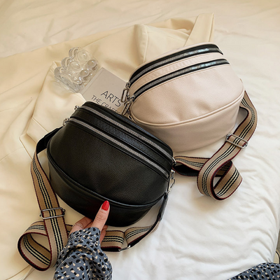 Deux sacs banane en cuir noir et beige avec fermetures éclair et bandoulières rayées, posés sur un lit blanc avec un livre et un verre à côté.