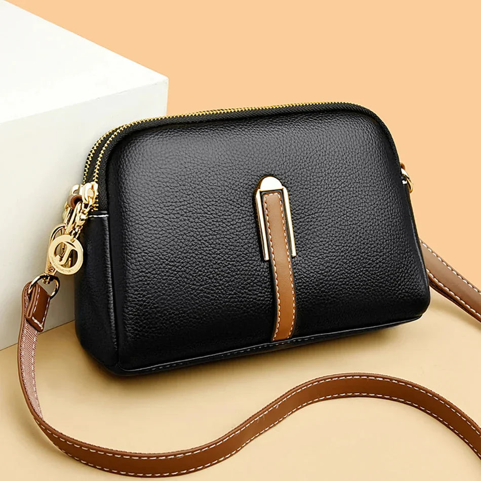 Sac à main en cuir noir élégant avec bandoulière marron, fermeture éclair dorée et détails métalliques, posé sur fond beige. Accessoire mode chic.