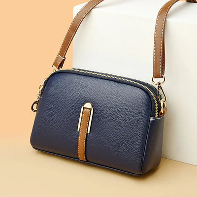 Sac à main en cuir bleu marine avec bandoulière marron, fermeture éclair dorée, posé sur fond beige. Accessoire mode élégant et tendance.