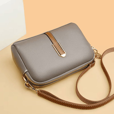 Sac à main en cuir gris avec bandoulière marron, fermeture éclair dorée et boucle décorative, posé sur fond beige. Accessoire mode élégant.