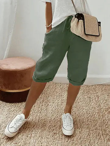 Femme portant un short vert kaki, t-shirt blanc, baskets blanches et sac à main en paille, posant sur un tapis beige. Mode décontractée chic.