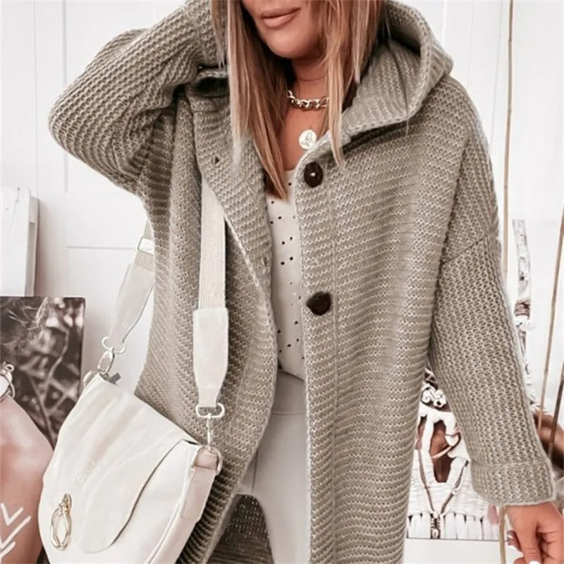 Femme portant un manteau en tricot gris avec capuche, sac à main blanc, collier doré, posant devant une photo. Mode automne-hiver tendance.