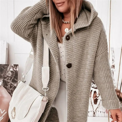 Femme portant un manteau en tricot gris avec capuche, sac à main blanc, collier doré, posant devant une photo. Mode automne-hiver tendance.