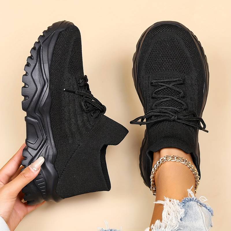 Chaussures de sport noires pour femme, semelle épaisse, design moderne, portées avec un jean déchiré, fond beige. Sneakers tendance et confortables.