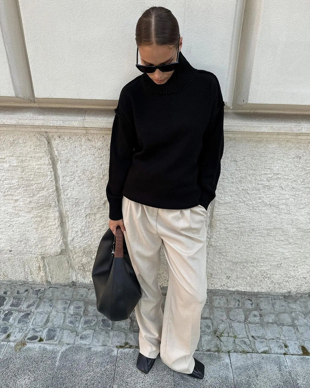 Femme en pull noir et pantalon beige, portant des lunettes de soleil et un sac noir, posant contre un mur en pierre. Mode automne chic et décontractée.