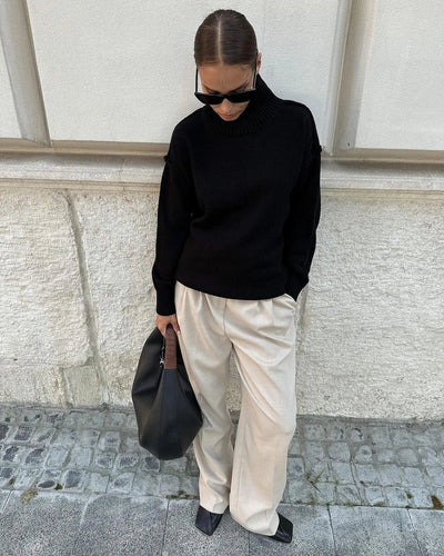 Femme en pull noir et pantalon beige, portant des lunettes de soleil et un sac noir, posant contre un mur en pierre. Mode automne chic et décontractée.