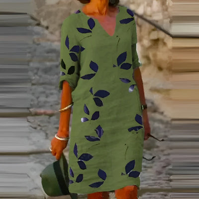 Robe verte à motifs de feuilles bleues, manches retroussées, portée par une femme tenant un chapeau. Mode estivale, style décontracté.