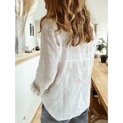 Femme de dos portant une chemise blanche en lin, cheveux ondulés, dans une cuisine moderne avec table en bois et plante verte. Style décontracté.