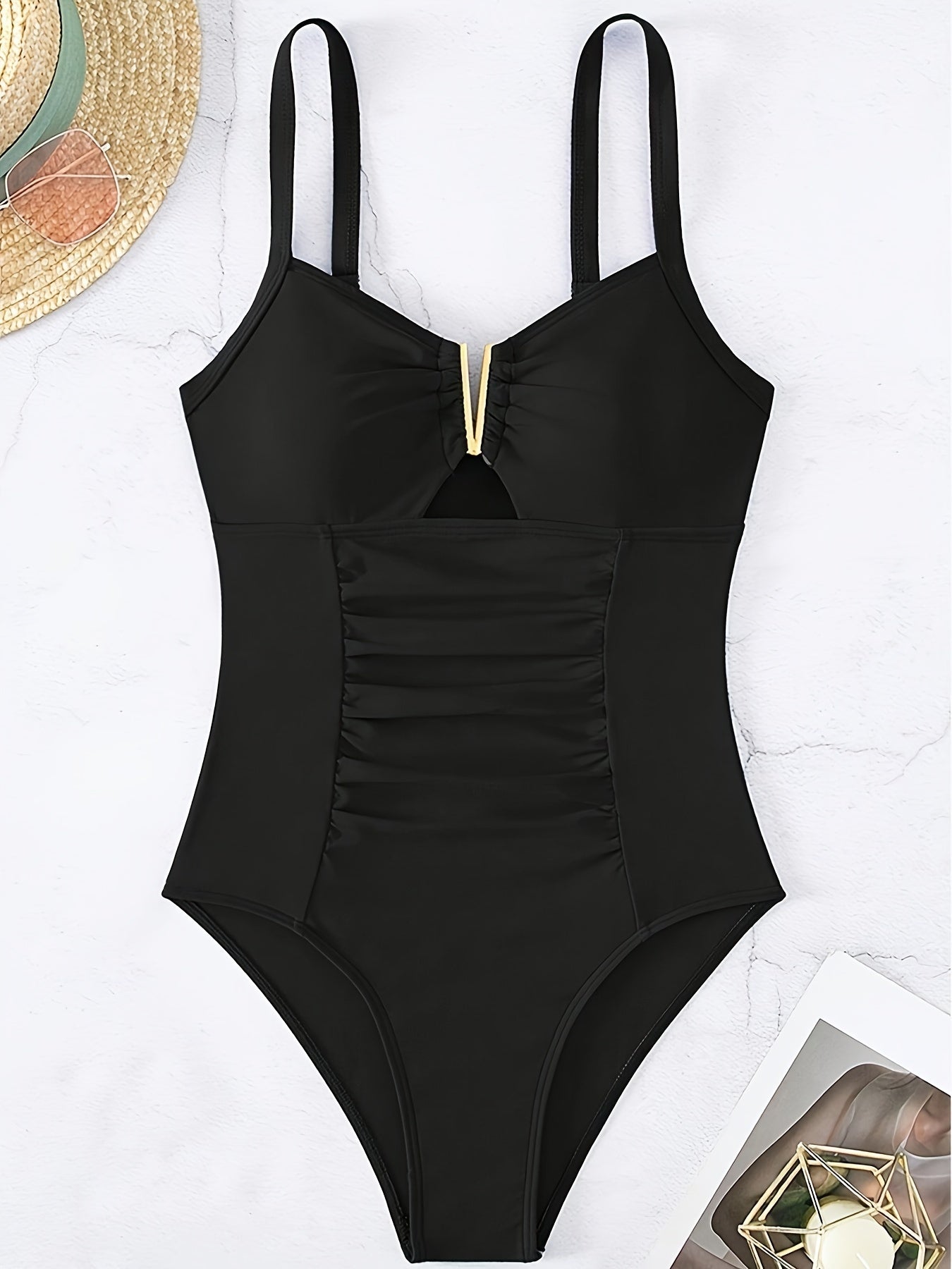 Maillot de bain une pièce noir élégant avec détails dorés, design froncé, posé sur fond clair avec chapeau de paille et magazine. Mode plage chic.
