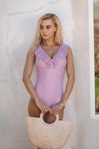 Femme en maillot de bain rayé rose et blanc, tenant un sac en osier. Mode plage, style estival, tenue élégante pour vacances en bord de mer.