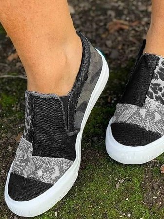 Chaussures slip-on noires et grises à motifs camouflage et serpent, semelle blanche, portées sur sol moussu. Mode décontractée, tendance urbaine.