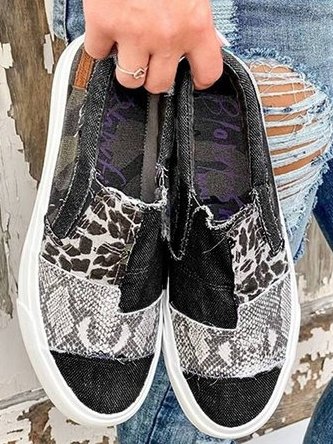 Chaussures slip-on en denim noir avec motifs léopard et serpent, semelle blanche, tenues par une main, devant un fond de bois vieilli et jean déchiré.