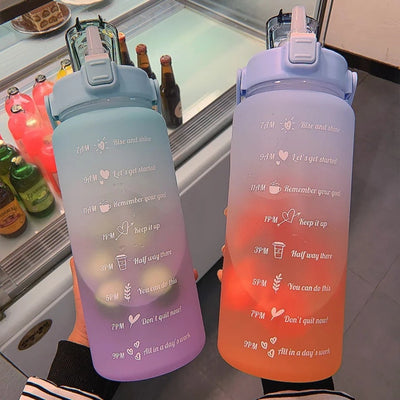 Deux grandes bouteilles d'eau graduées, dégradé de couleurs pastel, avec inscriptions horaires motivantes, posées devant un réfrigérateur.