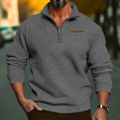 Homme portant un pull gris à col zippé, manches longues, sur fond urbain flou. Mode masculine automne, style décontracté, confort élégant.
