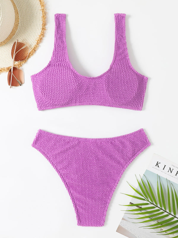 Bikini violet texturé sur fond blanc, accompagné d'un chapeau de paille, lunettes de soleil et magazine. Mode plage, maillot de bain tendance.