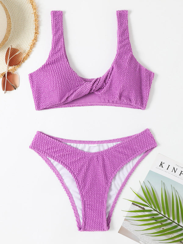 Maillot de bain bikini violet texturé, style deux pièces, avec chapeau de paille et lunettes de soleil, posé sur fond blanc avec magazine et feuille verte.