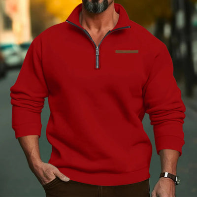 Homme portant un pull rouge à col zippé, manches longues, debout dans une rue floue. Mode masculine automne, style décontracté élégant.