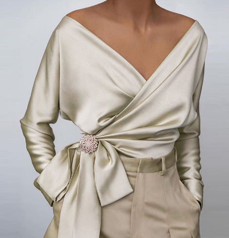 Blouse en satin beige élégante avec décolleté en V croisé, manches longues, ceinture nouée et broche décorative, associée à un pantalon assorti.