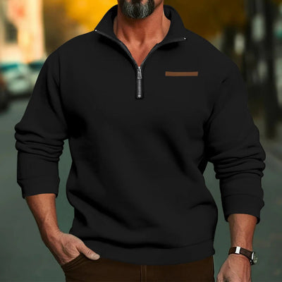 Homme portant un pull noir à col zippé, manches longues, sur fond urbain flou. Mode masculine, style décontracté, automne-hiver.
