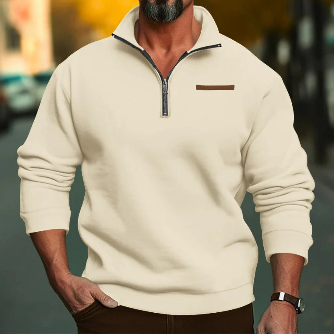 Homme portant un pull beige à col zippé, manches longues, debout dans une rue floue. Mode masculine automne, style décontracté élégant.