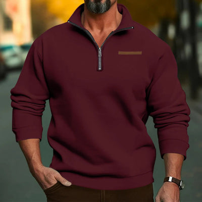 Homme portant un pull bordeaux à col zippé, manches longues, sur fond urbain flou. Mode masculine automne, style décontracté élégant.