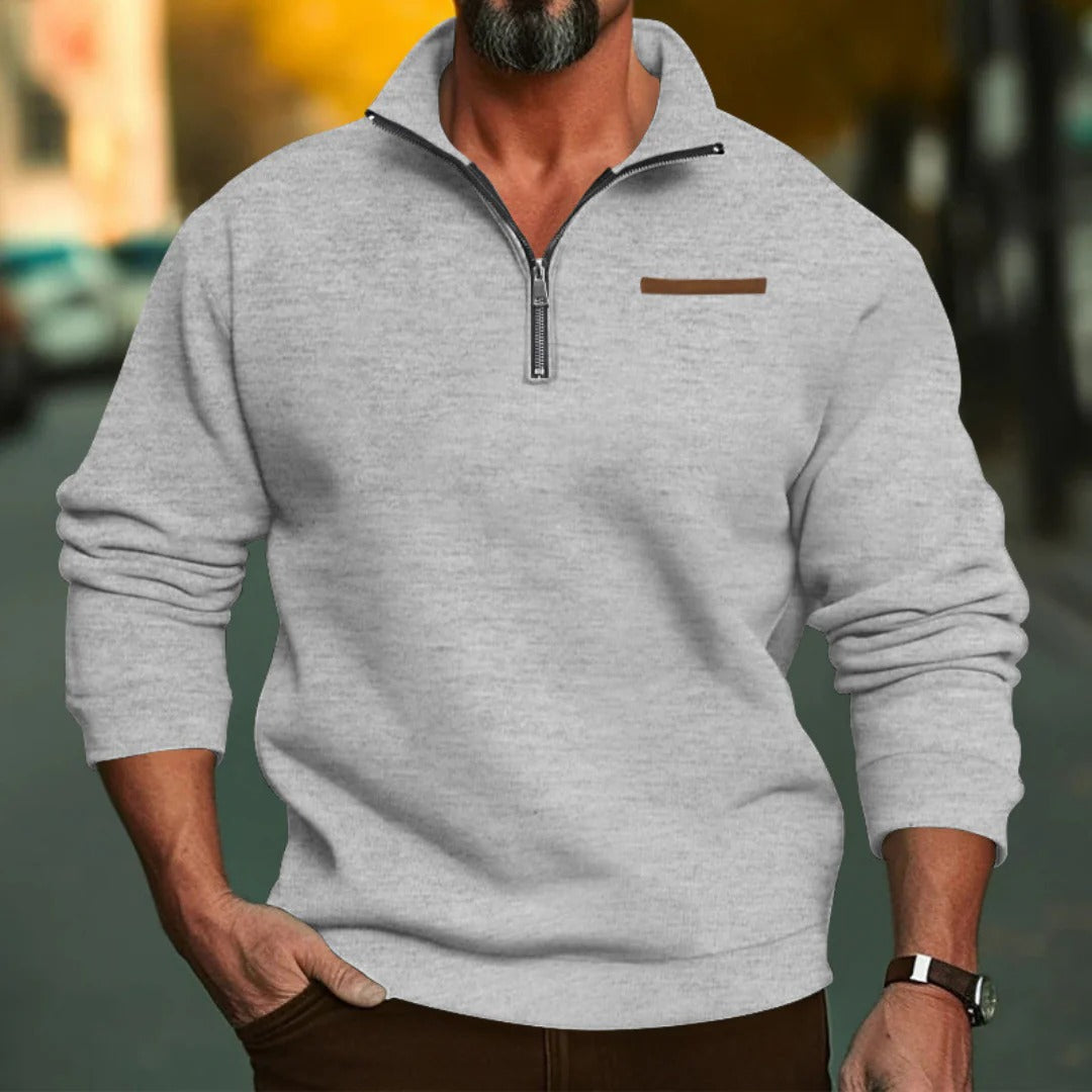 Homme portant un pull gris à col zippé, manches longues, sur fond urbain flou. Mode masculine automne, style décontracté, confort élégant.
