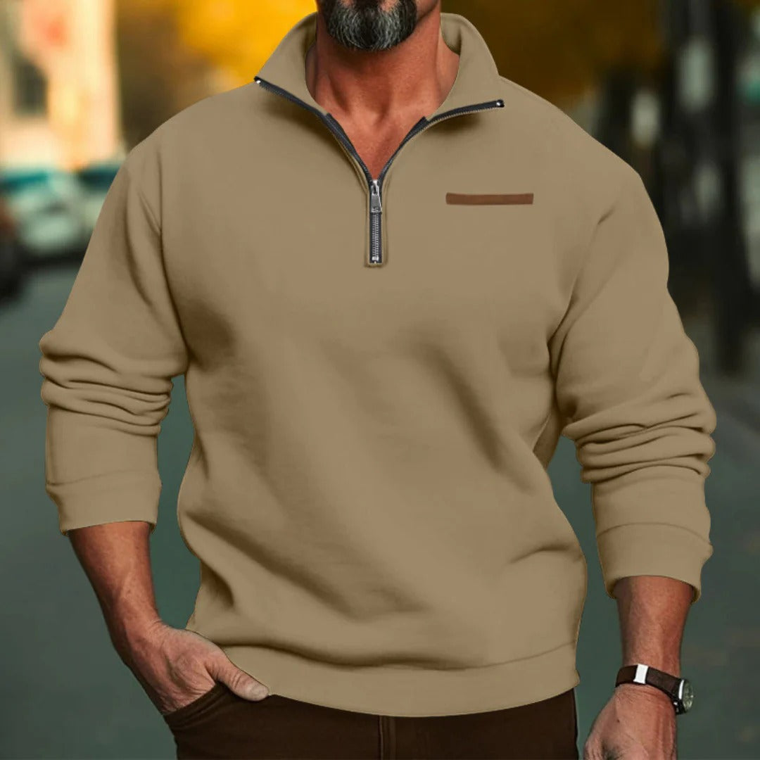 Homme portant un pull beige à col zippé, manches longues, sur fond urbain flou. Mode masculine automne, style décontracté, tendance actuelle.