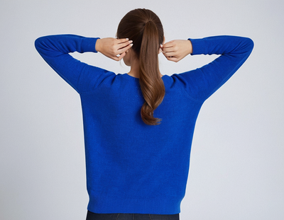Femme de dos portant un pull bleu vif, cheveux attachés en queue de cheval, posant les mains sur la tête. Mode automne-hiver, style décontracté.