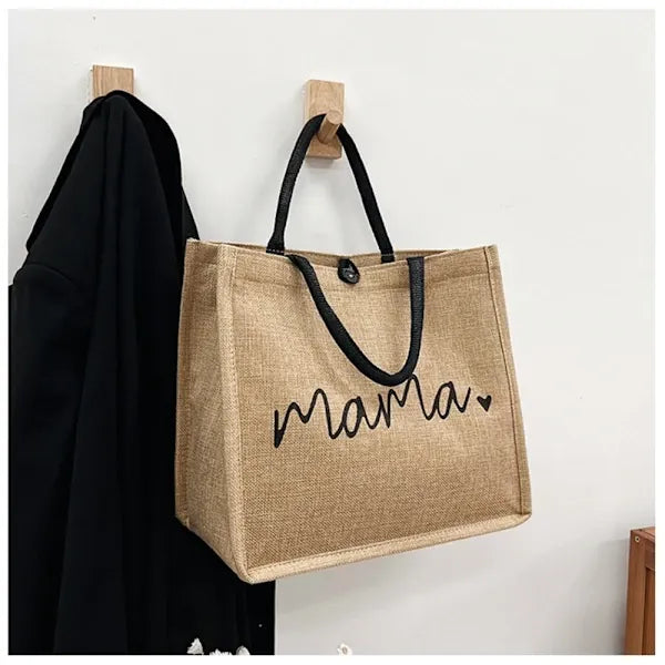 Sac en toile de jute beige avec poignées noires, inscription "mama" et cœur, accroché à un crochet mural. Accessoire mode écologique.