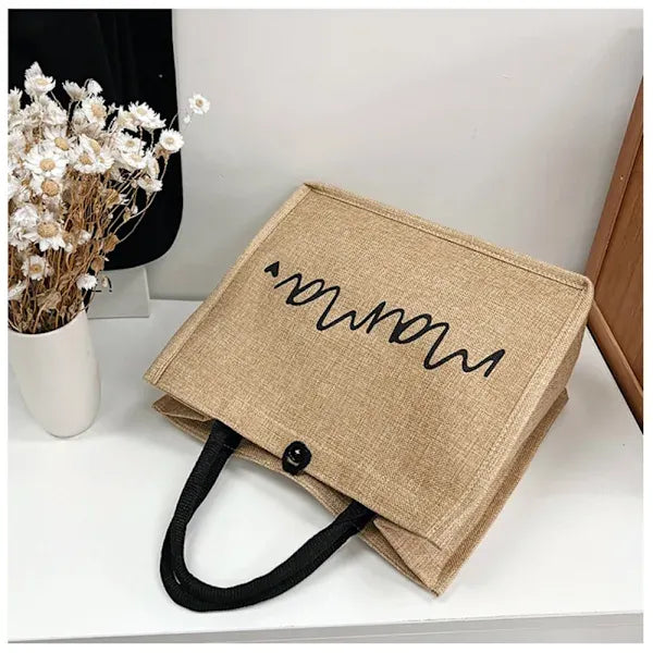 Sac en toile de jute beige avec motif noir ondulé, posé sur une table blanche à côté d'un vase de fleurs séchées. Accessoire mode écologique.