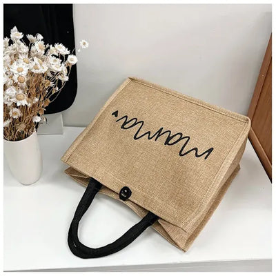 Sac en toile de jute beige avec motif noir ondulé, posé sur une table blanche à côté d'un vase de fleurs séchées. Accessoire mode écologique.