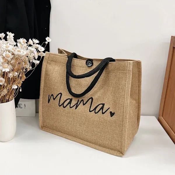 Sac en toile de jute avec inscription "mama", poignées noires, posé sur une table blanche à côté d'un vase de fleurs séchées.